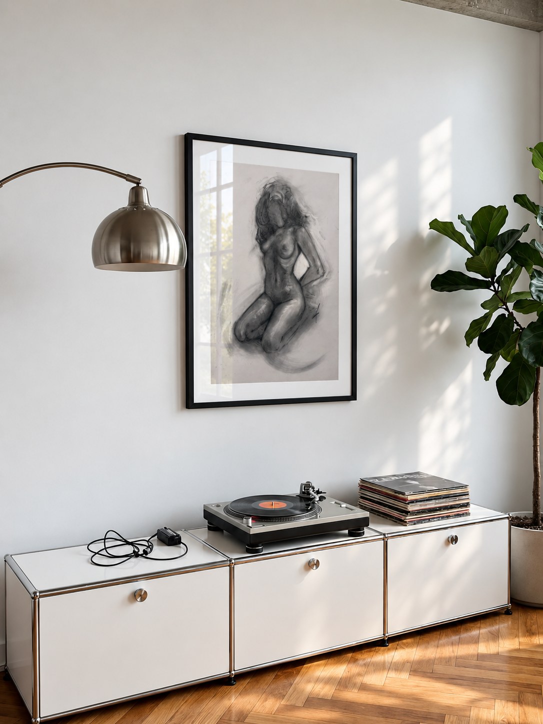 Charcoal drawing of a kneeling nude figure in a black frame, hung above a white modular sideboard with a turntable, beside an arc floor lamp and a fiddle-leaf fig, installation view
