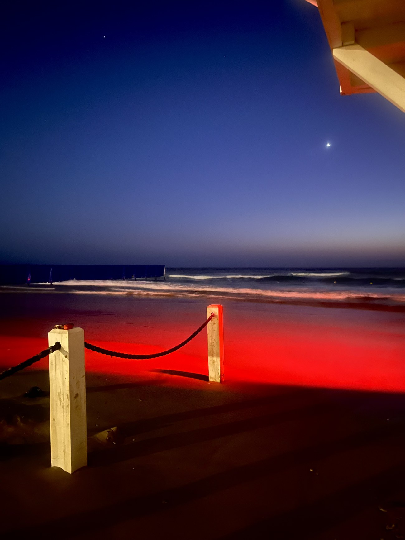A red-lit post and rope barrier on the sand at dusk, breakwater and waves beyond