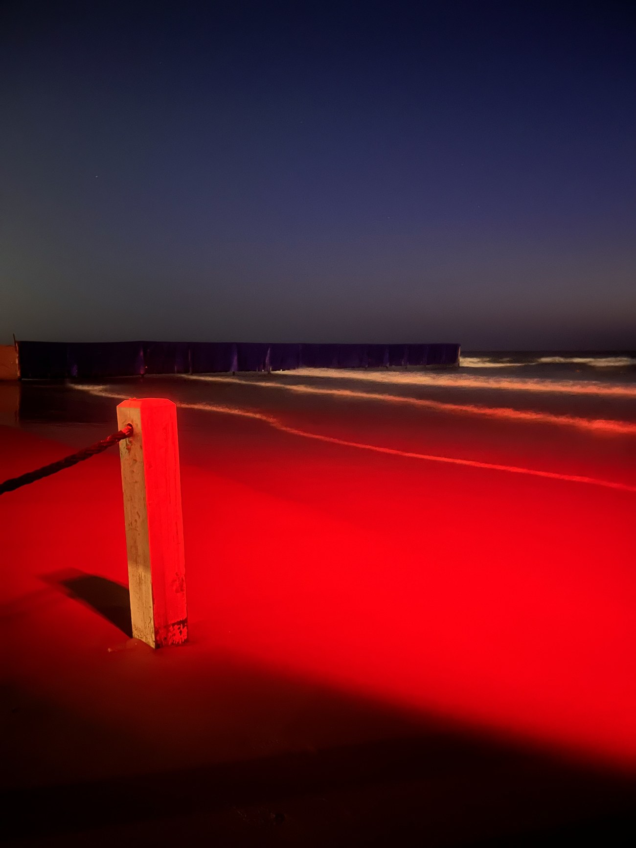 Close view of a wooden post glowing red under the neon sign, sand lit crimson