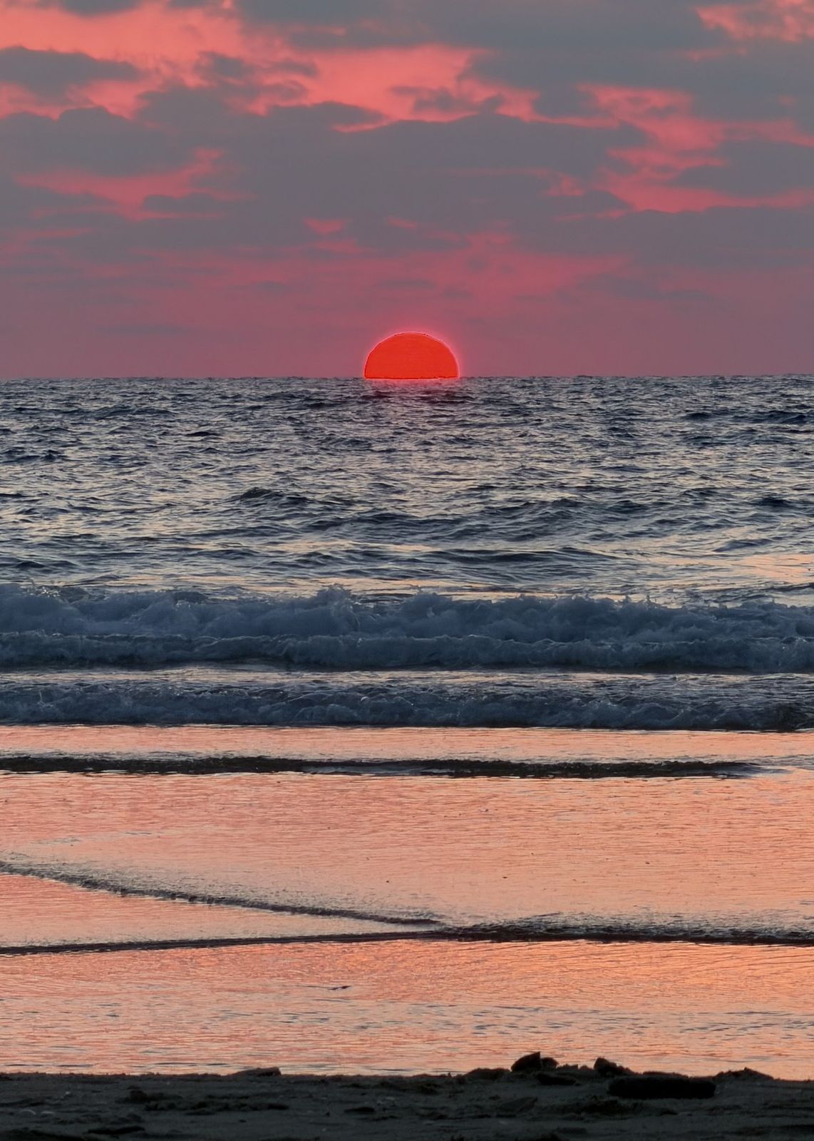 The sun setting into the sea, glowing red above the waves, pink clouds overhead