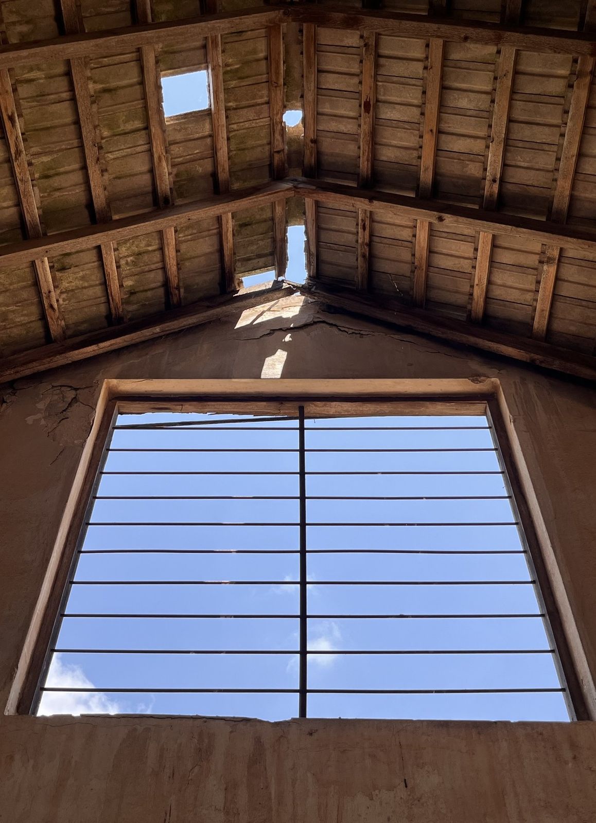 Looking up through the broken gable roof to a large barred window and sky