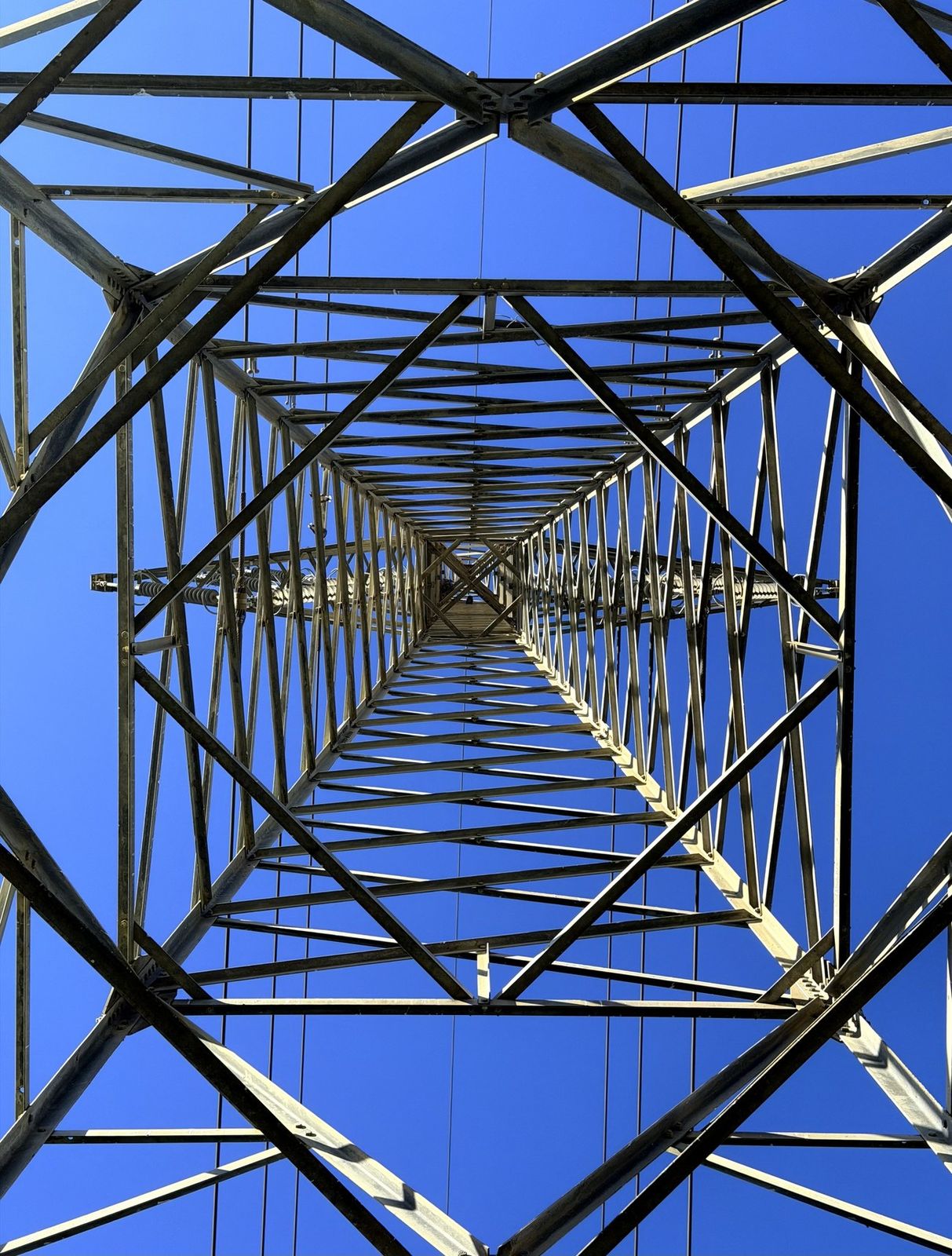 Looking straight up through a steel transmission tower, a symmetrical lattice against a blue sky