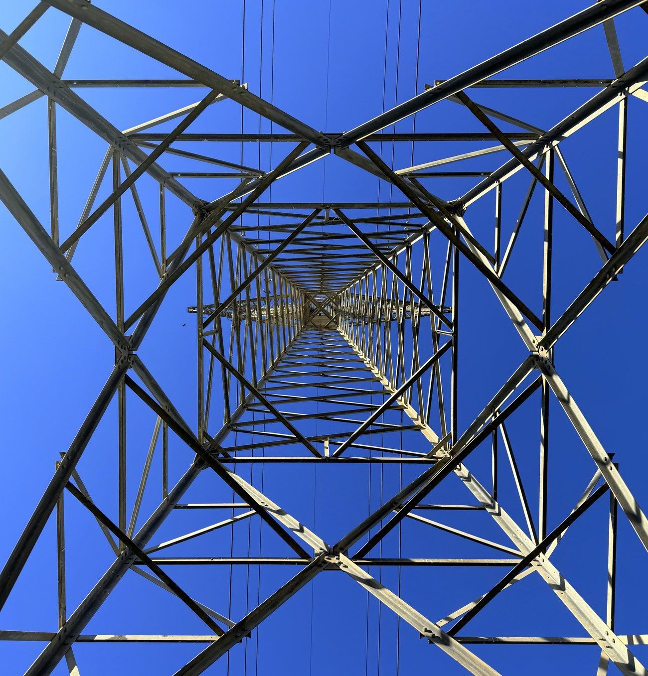 Looking straight up through a steel transmission tower, receding lattice against a blue sky