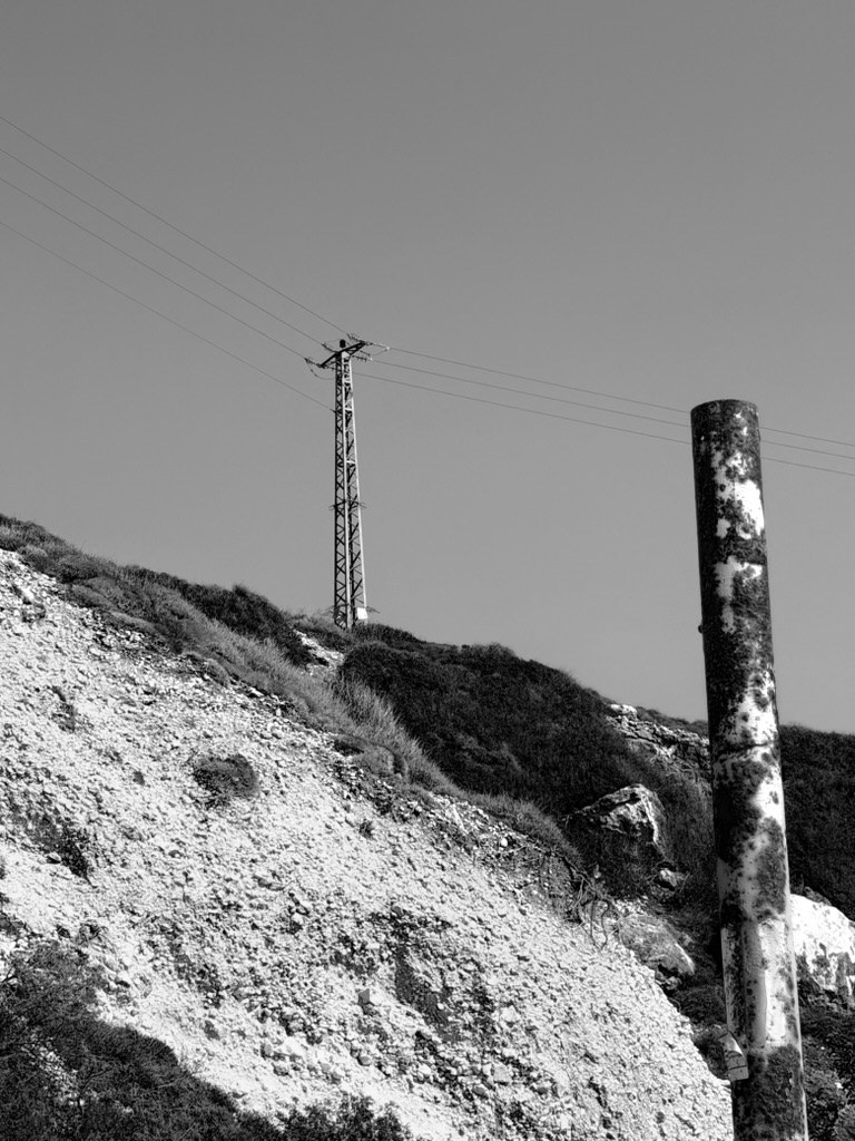 A steel power pylon on a scrubby hillside, a weathered, rust-streaked pole in the foreground, black and white