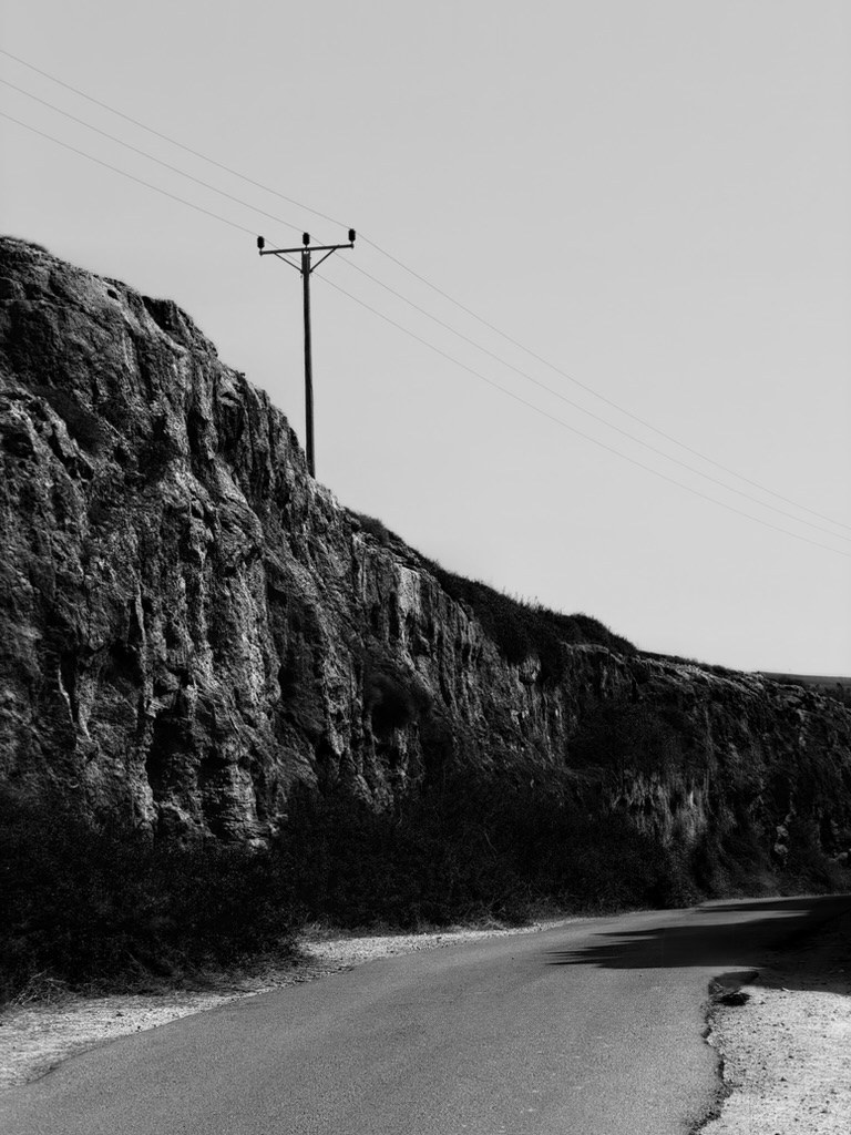 A single wooden power pole on top of a rough rock face above a narrow empty road