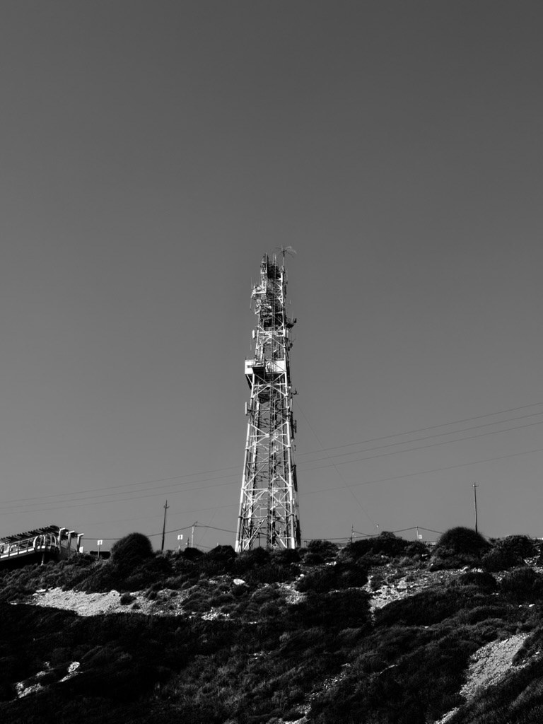 A tall lattice telecom tower on a hilltop covered in low scrub, against a flat dark sky