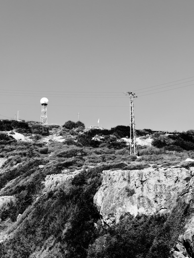 A white radar dome on a lattice tower and a power pylon on a rocky hill covered in scrub
