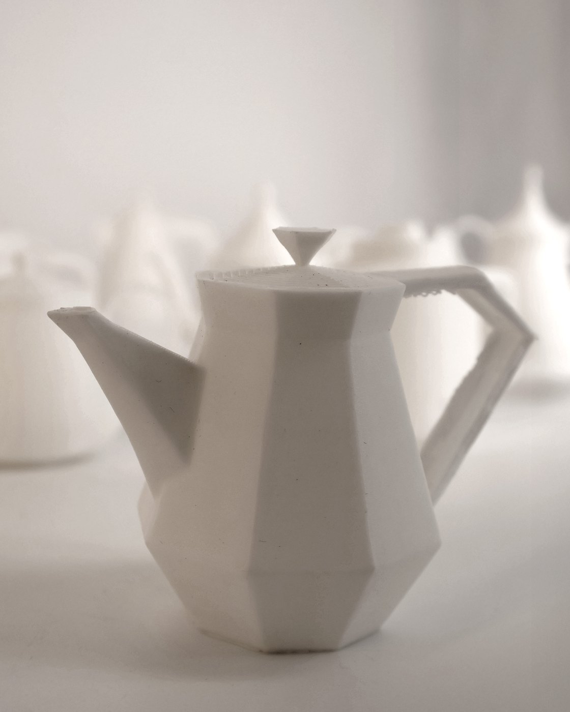 A white faceted 3D-printed teapot with a diamond-shaped lid knob, other printed studies blurred behind it